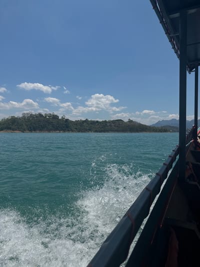From the perspective of a boat traveling on Cheow Lan Lake in Ban Ta Khun, Thailand, the image captures the surrounding environment. The boat's frame occupies portions of the top and right sides of the image. On the left, the sky is blue with scattered white clouds. In the center, the shoreline is visible, covered with dense green forest stretching towards the horizon. The lake itself is a deep turquoise. Water splashes up against the side of the boat, creating white foam and spray.