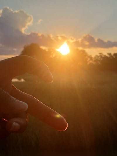 A hand, oriented vertically, points a single finger towards a bright light source, likely the sun. The sky is visible in the background, with clouds on the left side of the image. The location and city are unknown. The setting is an outdoor environment. The interaction between the hand and the sun, as perceived in the image, suggest an attempt to touch or point to the sun.