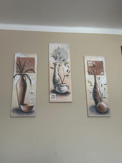Three vertical paintings are displayed on a neutral-colored wall. Each painting depicts a still life arrangement featuring pottery and foliage. The leftmost painting shows a tall, slender vase with long leaves, accompanied by a small bowl at its base. Two rectangular elements resembling stamps are positioned to the right of the vase. The middle painting features a gray vase with a smaller pot beneath it, from which delicate branches with leaves emerge. Similar stamp-like elements are present on this canvas. The rightmost painting displays a tall, curved vase with a rounded, bulbous object at its base, and branches with leaves extending upwards. This painting also includes the decorative stamp-like elements. The wall appears to be indoors, and the lighting suggests it is daytime. The paintings are hung in a row, with some space between them. The overall style of the artwork is consistent across all three pieces, suggesting they are part of a set. The location is identified as Teplice, Czechia.