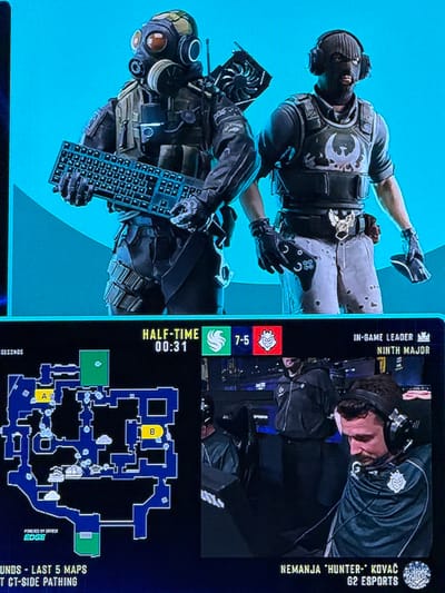 The image is a screenshot from a professional esports match, likely Counter-Strike: Global Offensive, at the DreamHack Masters Spring 2021 tournament held at the Prioritet Serneke Arena in Gothenburg, Sweden.

In the top panel, two figures are depicted in tactical gear. The figure on the left wears a gas mask and holds a keyboard. The figure on the right wears a balaclava and headphones, and appears to be holding a computer graphics card. Both figures are holding these items as if they are weapons, suggesting a humorous or thematic presentation.

The bottom panel shows a game interface. On the left, a minimap of a game level is displayed, with points of interest labeled 'A' and 'B'. The score "HALF-TIME 00:31" is visible, indicating the current game status. To the right of the minimap, player information is displayed. The score is shown as "7-5" with team logos. Below the score, the "IN-GAME LEADER NINTH MAJOR" is identified. Further to the right, a player named Nemanja "hunter-" Kovač from G2 ESPORTS is shown wearing headphones and a team jersey. He appears to be looking down, possibly in concentration or dejection.

The overall scene suggests a live broadcast or recording of an esports event, highlighting game elements and player status. The background, though not clearly visible, appears to be a stadium or arena setting.