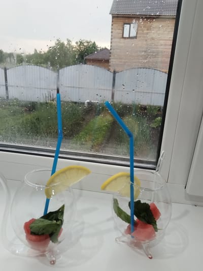The image depicts two clear glass teapots, each containing watermelon pieces and mint leaves, with a slice of lemon and a blue straw. Both teapots are positioned on a white windowsill, with a window behind them showing a rainy day scene outside. The view through the window includes a wooden house with a dark roof, a metal fence, and trees. The location is specified as Bezlesnyy, Russia.