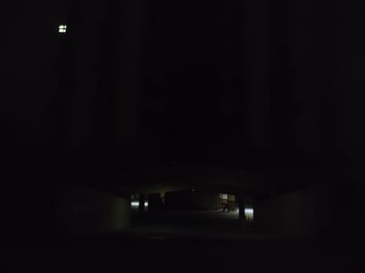 The image depicts an underground or enclosed area, possibly a parking garage or underpass, in Roranwala, Pakistan, at night. The scene is very dark with limited illumination. In the upper left quadrant of the image, a cluster of small, bright, rectangular lights is visible against the blackness. The central portion of the image shows the entrance to a darker area, with faint outlines of concrete structures and pillars. On the right side of this darker area, a motorcycle or scooter is parked near a wall with yellow and black striped markings. A single, bright light source is illuminating this area from the right. The overall impression is one of a dimly lit, possibly abandoned or underutilized urban space.