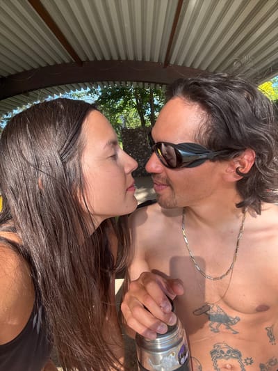 A man and a woman are angled toward each other, seemingly about to kiss or perhaps already kissing. The man is shirtless, wearing dark sunglasses, and holding a metal flask. He has dark hair pulled back, facial hair, tattoos on his chest and stomach, and a gold chain around his neck. The woman has long, dark hair. The setting appears to be outdoors, underneath a corrugated metal structure, with trees visible in the background. Without additional information, it is impossible to definitively say that the location is Langenfeld (Rheinland), Germany.