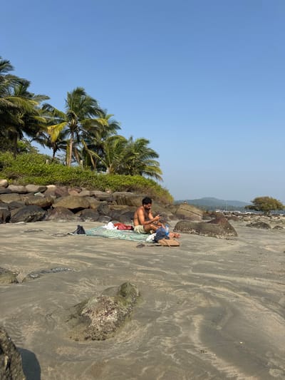On a beach, a man sits on a mat, near a rock barrier covered in vegetation. The beach is sandy, with a mix of light and dark shades, marked by patterns on the sand surface. The man appears to be using a phone. He is surrounded by personal items. Behind the rock barrier, there are tall palm trees reaching towards the clear blue sky. The horizon is visible in the distance, appearing slightly hazy.