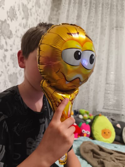A young person is partially obscured by a golden smiley-face balloon held in their right hand. The balloon has large, wide eyes and a distressed expression. The person is wearing a dark t-shirt with a graphic design. The background consists of a floral wallpaper and a white lace curtain. In the background, several stuffed toys are visible, including an avocado, a pink bird, and other plush toys. The scene appears to be indoors, possibly in a bedroom.