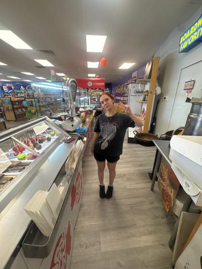 A person is standing in what appears to be a candy store. The person is wearing a dark t-shirt with a graphic on it and dark shorts. They are making a peace sign with their right hand. The store has shelves stocked with various items, a refrigerated display case filled with candy apples, and a checkout counter. In the background, there are signs and displays related to candy. One sign on the wall reads "EATALY." The lighting suggests it is daytime, and the scene appears to be indoors. The overall atmosphere seems casual and commercial, typical of a retail environment.