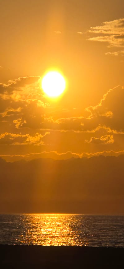 The image depicts a vibrant sunset over a body of water, likely the Issyk-Kul lake in Boz Beshik, Kyrgyzstan. The sun, a brilliant white orb, is partially obscured by scattered, golden clouds, casting a warm, orange glow across the sky. Rays of sunlight pierce through the atmosphere, creating a soft, diffused light. Below the sky, the water reflects the fiery hues of the sunset, creating a shimmering, golden pathway across its surface. The foreground is dominated by a dark, silhouetted shore, providing a stark contrast to the luminous sky and water. There are no people or visible objects other than natural elements. The environment is serene and tranquil, captured during the twilight hours.