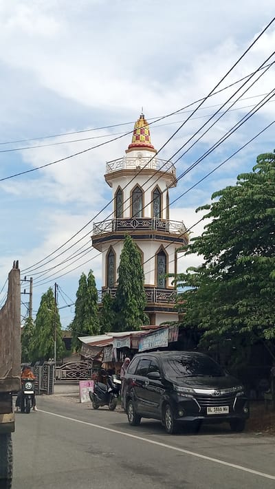 A bright, overcast day in Lhokdagang, Indonesia, features a prominent white minaret with ornate arched windows and a colorful tiled conical roof. The minaret is surrounded by lush green trees and is visible behind a row of small shops with corrugated metal roofs. In the foreground, a black SUV with a license plate reading "BL 1866 NH" is parked on the side of a paved road. To the left of the SUV, a black motorcycle is parked, and a person wearing a headscarf is standing nearby. Further down the road, another motorcycle with a rider wearing an orange jacket is visible. The scene is framed by numerous overhead power lines crisscrossing the sky.