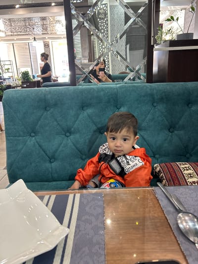 A young child wearing an orange jacket with white writing is seated on a teal tufted chair. There's a spoon and knife on a table with a grey patterned placemat. A white napkin is also on the table. The child is looking at the camera. Reflections in a decorative mirror show other people and the interior of a restaurant. The image was taken in Ақсу, Kazakhstan.