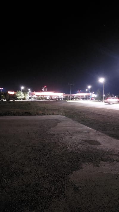 The image depicts an outdoor scene at night, likely near an airport in Kurylys, Kazakhstan. In the background, a brightly lit building, possibly an airport terminal, is visible with illuminated windows and signage. Several tall lampposts with bright lights line the area in front of the building and along a road. On the right side of the road, a vehicle's headlights and taillights are blurred due to motion, indicating it is driving. The foreground consists of a rough, dark, paved surface with some patches of discolored or wet areas. A grassy area with sparse vegetation separates the paved foreground from the road and building. The sky is dark, with a few faint stars visible.