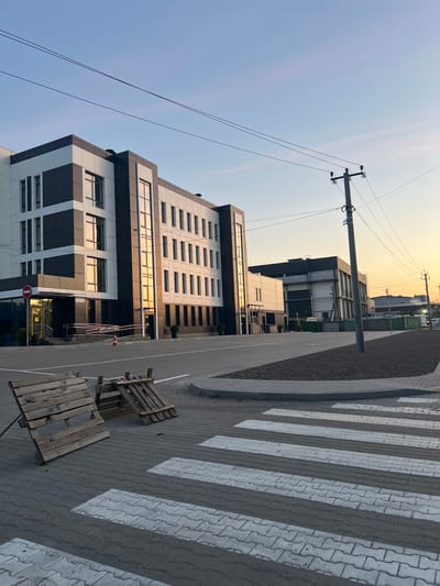 The image depicts a modern commercial building with a black and white facade, large windows reflecting the sunset sky, and a paved entrance with a ramp. In the foreground, a pedestrian crossing with white stripes is laid on a grey brick sidewalk. Two wooden pallets are placed on the left side of the pedestrian crossing. The scene is set against a clear sky transitioning from blue to orange at the horizon, with visible power lines and a utility pole. In the background, other buildings, fences, and a road are visible, suggesting an urban or industrial area. The location is identified as Horenychi, Ukraine. There are no people or animals visible in the image, and no discernible actions or interactions are taking place.