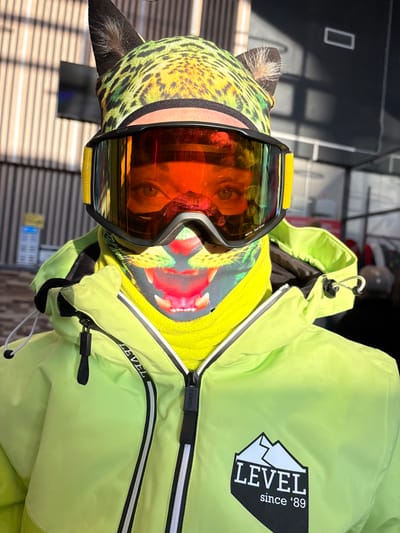 A person is depicted from the upper chest upwards, positioned outdoors in Korobitsyno, Russia, wearing winter sports attire. The individual's eyes are visible through large, black-framed ski goggles with highly reflective orange-red lenses, marked "NEON Red Iridium" on the lower right lens. A lime-green leopard-print beanie with dark, pointed animal ears covers the head. Below the goggles, a neck gaiter or balaclava with a graphic print of a roaring big cat's open mouth and fangs is visible. The person wears a bright neon-lime green winter jacket, zipped up, revealing a yellow inner layer. The jacket features a black and white logo on the right chest depicting a mountain silhouette above "LEVEL" and "since '89" below it. A black zipper pull with "LEVEL" embossed is visible. The background shows a vertical slatted building facade, indicative of a ski resort or outdoor recreational area, under direct sunlight.