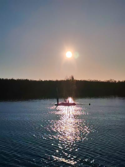 A landscape photograph captures a body of water, likely a lake or wide river, at sunset. The sun, a bright white orb, dominates the sky, positioned high above the horizon and reflecting intensely off the water's surface.
A small, isolated island sits in the middle of the water, supporting a few trees and vegetation. The sun casts a shimmering reflection that stretches across the water from the island towards the viewer. A dark line of dense forest runs along the distant horizon.
The color palette is dominated by the warm tones of the setting sun, with hues of orange, yellow, and hints of pink in the sky. The water appears dark blue, contrasting with the sun's reflection and the illuminated areas around the island. The location is in Kurdiug, Russia.
