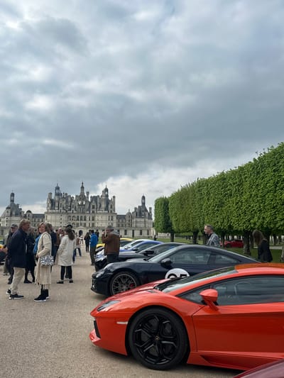 The image shows a group of people standing in front of a row of luxury sports cars parked outside the Chateau de Chambord, located in Chambord, France (latitude: 47.615, longitude: 1.519). The people are dressed casually and appear to be admiring the cars. The cars are all high-end models, including a Lamborghini Aventador, a Porsche 911, and a Ferrari. The chateau can be seen in the background, towering over the scene. The sky is overcast with clouds, and the trees are in full bloom, creating a lush and picturesque setting.