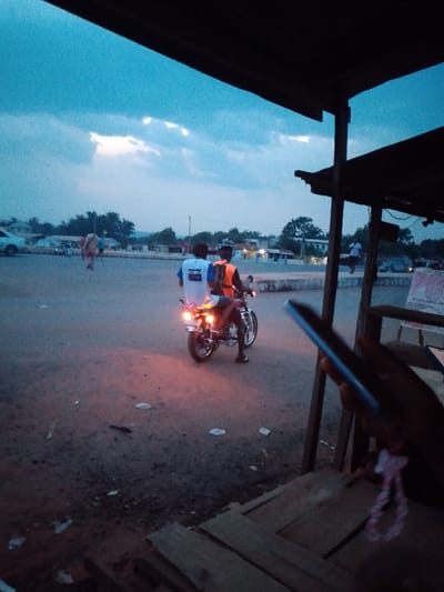 The image, likely taken in Urualla, Nigeria, depicts a scene at dusk. The sky is a deep blue with scattered clouds. The foreground shows a dirt ground littered with paper debris. In the middle ground, two people are on a motorcycle with the headlights on. The passenger in the back wears a white shirt with blue design. The driver wears a bright orange vest. In the right foreground, there is a structure with wooden posts and a roof, partially obscuring the view. A hand holding a mobile phone is visible in the lower right corner. In the background, there are parked cars, people walking, and buildings under a darkening sky.
