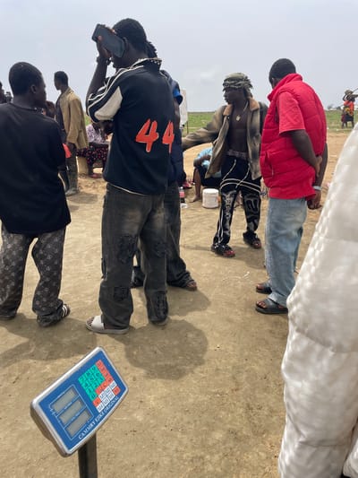 The image shows a group of people standing on a flat, sandy surface, possibly in Maiyanga, Nigeria. Visible are a digital scale, legs and torsos of individuals in various clothing styles, including a red puffer jacket, black shirts, patterned pants, and ripped jeans. One person is holding a phone. The background suggests an open, outdoor environment.