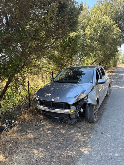 The image shows a heavily damaged silver sedan on the side of a paved road. The front of the car is significantly crushed, with the bumper missing and internal engine components exposed. The hood is dented and crumpled, and the headlights appear to be intact. The driver's side front wheel is turned outwards. The car is partially off the road, with its left side near a wire fence and dense vegetation. Trees with green foliage form a canopy over the scene, with dappled sunlight filtering through. The road itself appears to be in a rural or semi-rural setting. There are no people visible in the image. The context of Santo-Pietro-di-Tenda, France is not visually evident within the image itself.