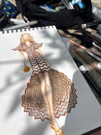 A fashion sketch of a woman in a zigzag patterned dress is depicted on a white sketchbook page. The sketch is done with colored pencils and markers. The woman has blonde curly hair and is holding a round, yellow object in her left hand. Her dress has a V-neck and short sleeves, with a flared, floor-length skirt. Orange sandals are visible on her feet.
The sketchbook is open on a textured surface, possibly concrete or stone, with dappled sunlight casting shadows. Surrounding the sketchbook are various art supplies, including colored pencils, markers, and a black fabric pouch. Some of the markers are Promarker Brush pens, and one is labeled with "G94" and "Olive". The art supplies are scattered around the sketchbook, suggesting an outdoor creative session. The image does not provide any specific information about the location in the city of O Casal, Spain.