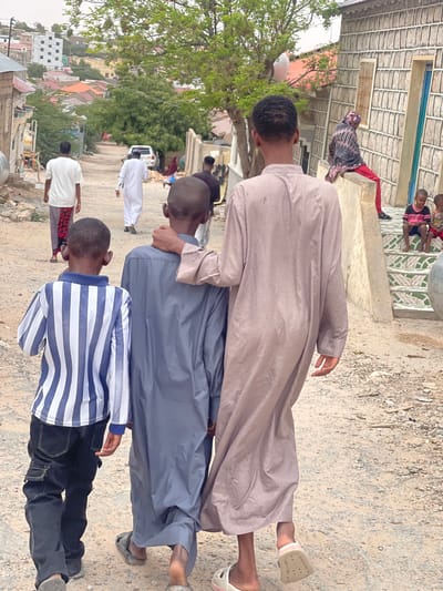 In Hargeisa, Somaliland, a group of boys walks down a dusty, unpaved street lined with buildings. The architecture suggests a blend of traditional and modern styles, with some structures appearing to be made of concrete and corrugated metal, while others are more elaborately designed with stone facades. The road is uneven and stony, indicative of unpaved urban infrastructure common in many parts of the city.
In the foreground, three boys are walking away from the camera. The boy on the left is wearing a blue and white striped long-sleeved shirt and dark cargo pants. The middle boy is in a dark blue thobe. The boy on the right, the tallest of the three, is wearing a light brown thobe. He has his arm around the shoulders of the middle boy. Further down the street, two other boys are visible walking towards the camera, one in a white thobe and the other in a white t-shirt and patterned pants. In the background, more people are seen, including a woman in a colorful headscarf and red pants sitting on a low wall, and two young children sitting on steps. A white SUV is parked on the side of the road. The surrounding environment is characterized by residential buildings, some with multi-story designs, and sparse vegetation, typical of an urban setting in a semi-arid climate. The overall impression is of a community going about its daily life in a bustling, developing urban area.