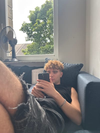 The media file shows a young person lying on a sofa, holding a smartphone. The person has curly, light brown hair and is wearing a black t-shirt and dark denim shorts. A silver bracelet is visible on their left wrist. The scene appears to be indoors, with a window to the left revealing trees and a building outside, suggesting a daytime setting. Natural light streams in from the window. A black fan is positioned on a windowsill below the window. The sofa is dark blue with a patterned cushion. The overall mood seems relaxed and casual. There is no visible text in the image. The location context provided is Hamm, Germany.