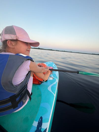 In this media file, a young girl wearing a pink baseball cap and a life vest lies on a paddleboard. The paddleboard is predominantly teal with a blue wave pattern and a dark blue shark graphic. She is holding a paddle with a green and white blade. The girl is looking up and to the side with a serene expression. Her feet are bare and resting on the paddleboard. An orange object, possibly a bag or personal flotation device, is visible behind her.

The scene is set on dark, calm water with subtle ripples reflecting the sky. The sky is a pale blue with a hint of yellow on the horizon, suggesting it might be early morning or late evening. The lighting is soft, and the overall atmosphere is peaceful. The location is identified as Salaspils, Latvia. There is no visible text in the image.
