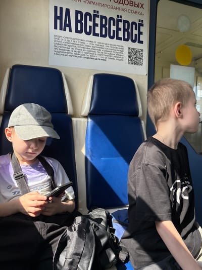 This image depicts the interior of a public transport vehicle, likely a train or intercity bus, within the city of Vyatskiye Polyany, Russia. Two pre-teen males are seated on blue and white upholstered seats.

On the left, a boy wearing a grey baseball cap with "NOFEAR" embroidered on the front, a light purple and white short-sleeved shirt, and dark pants, is seated with his head bowed, engrossed in a black smartphone held with both hands. A black strap from a bag or backpack crosses his chest. Strong sunlight illuminates his hands and the right side of his face.

On the right, a second boy with short, light brown hair and wearing a black short-sleeved t-shirt with white graphics on the front, is seated facing away from the camera, looking towards a window on his right. A thin silver chain is visible around his neck. His right arm is illuminated by sunlight.

Between the two boys, on the dark floor, a black backpack and a blue bag are visible. Above the seats, a white poster with Cyrillic text and a QR code is affixed to the wall. The poster advertises a "SUPERSMART PROMO" credit from "POCHTA BANK," detailing loan amounts, interest rates, and terms. The interior walls are light-colored above the seats. A window on the far right displays a circular yellow sticker and a rectangular white sticker. Intense sunlight streams from the right side of the frame, casting sharp shadows across the seats, the floor, and parts of the boys' attire.