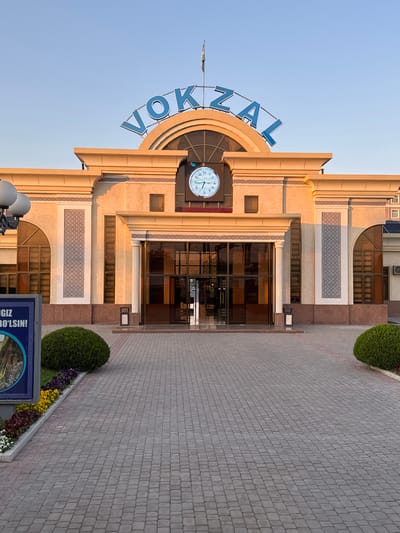 The image depicts the facade of the Jizzakh railway station in Uzbekistan. The building features a clock tower with a large analog clock displaying the time. Above the clock, the Cyrillic letters "VOKZAL" are visible, spelling out "вокзал," which means "railway station" in Russian. The architectural style appears to be a mix of Soviet-era and Central Asian influences. The entrance is marked by glass doors and decorative elements. The surrounding area includes paved walkways and ornamental landscaping with flowers and shrubs. A blue sign with Cyrillic text is visible near the bottom of the frame. The overall lighting suggests it is either early morning or late afternoon.