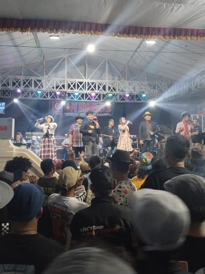 This is a medium shot of a live music performance on a stage under a large tent structure. The stage has a dark backdrop with a banner that reads "LOM LORENSA DANGDUT JA... INDONESIA." There are multiple performers on stage, including a woman in the center in a white shirt and a plaid skirt, holding a microphone and singing. To her right is a man in a fedora and a dark jacket, also holding a microphone. Other band members are visible playing instruments and singing. The stage is lit with bright spotlights and colored lights, creating a vibrant atmosphere. In the foreground, a crowd of people are watching the performance. Many in the crowd are wearing hats and casual clothing. The environment appears to be an outdoor event, likely in a public space in the city of Celep, Indonesia.
