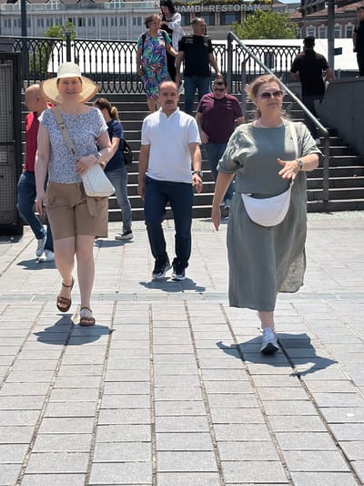In the city of Fatih, Turkey, a sunny day is captured on a cobblestone walkway. A group of people are walking on the walkway, with some on stairs in the background. A woman in the foreground on the left wears a floral print blouse, tan shorts, and a wide-brimmed straw hat. She carries a tan crossbody bag and a white purse. Beside her, a man in a white polo shirt walks. To the right, another woman walks in a long, olive-green dress with a white fanny pack. She wears sunglasses and a watch. Behind them, more individuals are seen walking in various directions and on stairs. The background shows buildings and a railing.