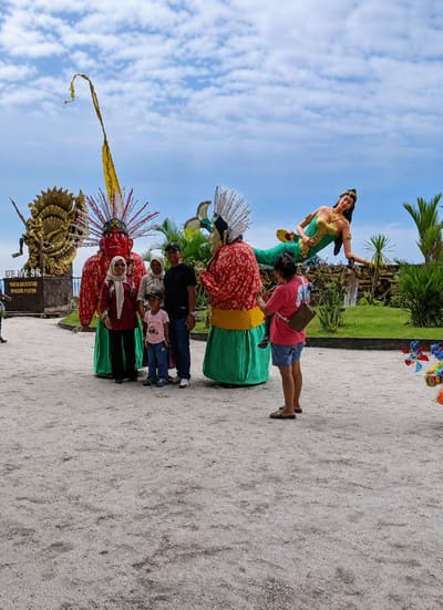 A family of four, including two adults, one child, and a baby, are posing for a photograph in front of two giant ogre statues in the city of Kota Pari, Indonesia. The father is wearing a black t-shirt and a dark baseball cap, and the mother is wearing a maroon headscarf and a maroon long-sleeved shirt with black pants. Their son is wearing a pink t-shirt and blue jeans, and the baby is being held by the woman on the right. The ogre statues are dressed in red and green traditional clothing. In the background, a statue of a mermaid can be seen. The ground is covered in sand, and palm trees and other greenery are visible. The sky is blue with white clouds.
