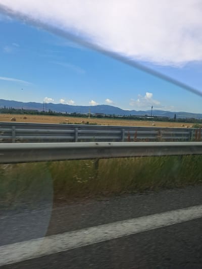 The image captures a view from a moving vehicle, likely on a highway, in Gorni Bogrov, Bulgaria. The perspective is from the side window, looking outwards. The scene is a landscape dominated by a clear blue sky with scattered white clouds on the left. A large, wispy cloud formation stretches across the upper left portion of the frame, creating a dramatic contrast with the bright sky.
To the right of the sky, a field of harvested crops, appearing as golden stubble, stretches horizontally. Several round hay bales are visible in the field, indicating it's likely late summer or early autumn. Beyond the field, a range of mountains forms the horizon, with some trees and structures, including power lines and what appear to be farm buildings, dotting the foothills.
In the foreground, a metal guardrail runs parallel to the direction of travel, separating the vehicle from the field. The road itself is visible on the far right of the image, with a white line marking its edge or a lane divider. The asphalt appears dark and slightly wet in places, suggesting recent rain or morning dew. The overall lighting suggests it is daytime, likely morning or afternoon, with the sun casting a bright glow. There are no people or animals visible in the frame. The mood is peaceful and suggests a journey through a rural area.