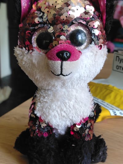 A close-up image depicts a plush toy, likely a fox or similar animal, sitting on a light wooden surface. The toy is characterized by its head and upper body covered in iridescent sequins that alternate between pink, silver, and black, creating a shimmering effect. It has a soft, white furry muzzle with a bright pink nose and a small black embroidered smile. Two large, prominent black eyes with sparkling silver rims give the toy a wide-eyed, endearing expression. The lower part of its body and its limbs are made of a fluffy black fabric.
The background is softly blurred, showing hints of an indoor environment. To the right, a vibrant yellow object, possibly a package or bag, is visible. In the upper right corner of the background, partial text in Cyrillic script can be seen on a white surface. The visible fragments read "Пу" and "ОТК," which likely correspond to words such as "Пункт" (point) and "Открыто" (open), aligning with the provided location context of Tserovo, Bulgaria. The lighting is soft and even, highlighting the reflective nature of the sequins. The toy appears to be stationary, presented as the main subject of the photograph.