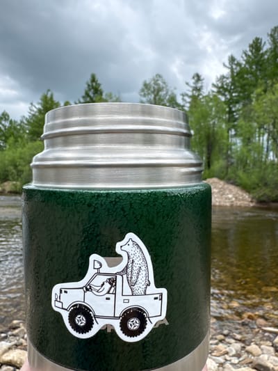 A close-up shot focuses on the textured, dark green body of a thermos. Adorning the thermos is a black and white sticker depicting a stylized vehicle, possibly a jeep, with a bear driving and another bear in the passenger seat. The thermos has a metallic silver top with a ribbed design. The background is out of focus, showing a body of water with rocky shores and lush green trees under a cloudy, grey sky. The overall scene suggests an outdoor, possibly natural, setting. The specific location of Муртыгит, Russia, cannot be determined from the visual information provided in the image.