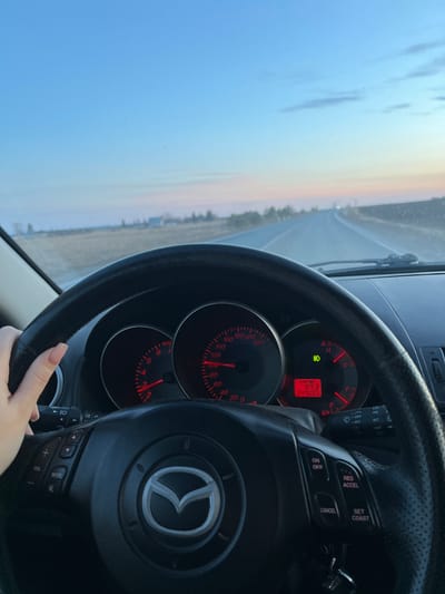 The image depicts the interior of a Mazda car, with the dashboard and steering wheel prominently visible. The odometer reads "347655" and the trip shows "97.3". The landscape visible through the windshield includes a road stretching into the distance, a field, some trees and a light sky, presumably captured near sunset or sunrise. The steering wheel is black with the Mazda logo in the center and features various buttons, including cruise control options. A hand with manicured nails is visible near the bottom of the steering wheel. Based on the environment, the image could potentially be taken near Kliuchi, Russia due to the flat landscape, however, without additional details, it's impossible to confirm the precise location.