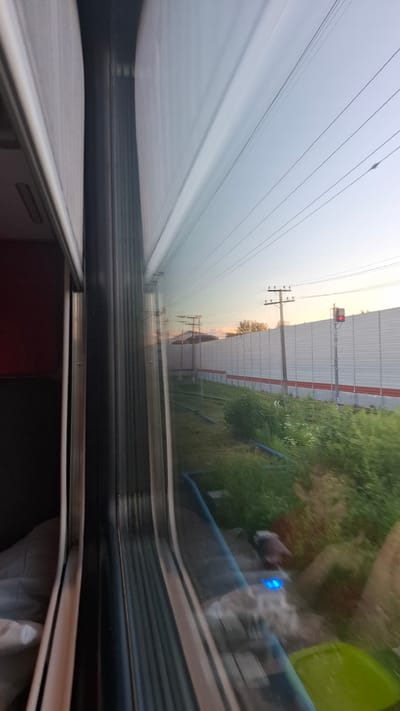 This media file captures a view from a moving train, likely in Volkhov, Russia, during a hazy twilight. The scene is dominated by a vast expanse of pale, overcast sky, crisscrossed by numerous power lines that extend diagonally across the frame. In the mid-ground, a tall, light-colored sound barrier runs parallel to the train tracks, punctuated by red stripes. A solitary traffic signal with a red light is visible atop one of the poles supporting the power lines. Beyond the barrier, lush green foliage and trees suggest a rural or semi-rural setting. The train's interior is partially visible in the foreground, showing a dark material and a metallic frame, suggesting the window of a passenger car. A faint blue light is visible within the train, possibly from a device. The overall impression is one of quiet, transient travel, with the subdued light and motion creating a somewhat melancholic or contemplative atmosphere.