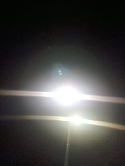 An intense, bright light source dominates a dark, seemingly nighttime scene. Lens flares streak horizontally and vertically, obscuring details. The darkness surrounding the light is nearly absolute, with minimal texture visible. The extreme contrast makes it challenging to discern the source or context of the light itself.