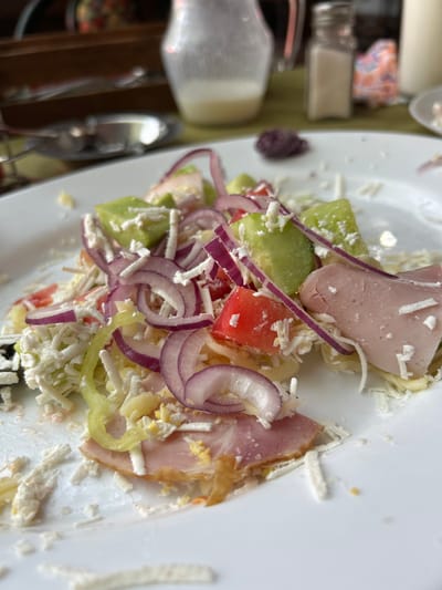 This is a close-up, overhead shot of a salad on a white plate. The salad appears to be a mix of chopped tomatoes, cucumbers, red onions, and cheese, with slices of ham visible. Some shredded cheese is also scattered around the plate. In the background, a blurry pitcher, a salt shaker, and a blurred object with a floral pattern can be seen. The setting appears to be a restaurant or dining area.