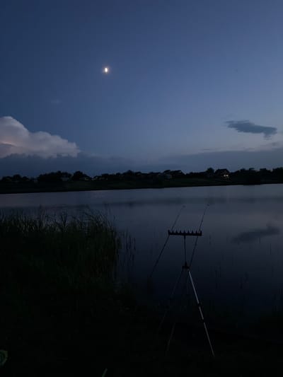 The image depicts a twilight scene at a body of water, likely a lake or pond, in Hrebinky, Ukraine. The sky is a deep blue, transitioning from a lighter shade near the horizon to a darker hue overhead. A crescent moon is visible in the upper left quadrant of the sky, accompanied by some scattered clouds. A large, illuminated cloud formation is present towards the lower left, hinting at possible rain or storm activity in the distance.

Along the shoreline, a silhouette of trees and houses can be seen, with one house in the middle ground featuring illuminated windows. This suggests a rural or semi-rural setting. In the foreground, on the right side of the image, a fishing rod holder with two fishing rods is set up, indicating that fishing is taking place. The water surface reflects the dim light of the sky and the surrounding landscape, with subtle ripples breaking the stillness. Tall grasses or reeds line the bank in the foreground, further defining the shoreline. The overall mood is tranquil and serene, characteristic of dusk by the water. There is no visible text in the image.