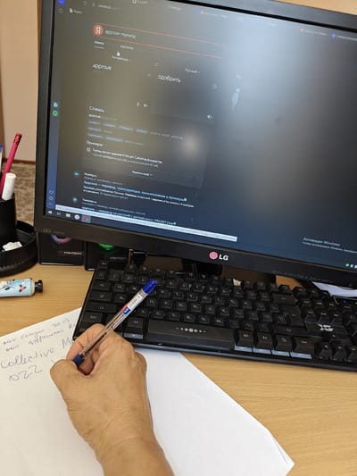 A person's hand, holding a blue pen, is writing on a white sheet of paper. The paper has some handwritten text in what appears to be Cyrillic script, including the words "Collective M" and "022". The person is seated at a wooden desk in front of a computer monitor. The monitor displays a Yandex Translate webpage with the English word "approve" being translated to Russian. The monitor also shows various application icons and browser tabs. A black keyboard is visible on the desk, along with a pen holder containing colored pencils. The overall environment suggests a home office or study space. The location in the city of Каракастек, Kazakhstan is not explicitly depicted in the image, but the Cyrillic script on the paper and the use of Yandex Translate suggest a connection to a Russian-speaking or Central Asian region.