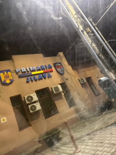The image displays the exterior of the "Primaria Jilava" (Jilava City Hall) building at night in Jilava, Romania. The building features a light-brown facade with a prominent illuminated sign bearing the words "PRIMARIA JILAVA" in blue and red lettering, incorporating the blue, yellow, and red colors of the Romanian flag in the "PRIMARIA" section. Two coats of arms, one on the left and one on the right, are affixed to the facade, flanking the main sign. Below the sign, three white exterior air conditioning units are mounted: two stacked vertically on the left side, and one single unit on the right. Several dark, rectangular windows are visible across the facade. A small yellow square sign with a black lightning bolt symbol is located on the lower left portion of the wall.
The ground in the foreground is paved with light-colored, possibly interlocking, pavers. A large, dark green waste bin with a lighter lid is positioned on the right side of the frame. Towards the bottom-center, a circular planter contains visible greenery, and a red pole or barrier stands vertically. Overhead, a utility pole heavily wrapped with numerous cables and wires extends diagonally from the upper right of the frame, supported by metal structural elements. The scene is illuminated by artificial light sources, primarily from the building's signage. The image displays reflections and streaks consistent with being taken through a glass surface, such as a window, and has a slight diagonal tilt to the right. No individuals are present in the scene.