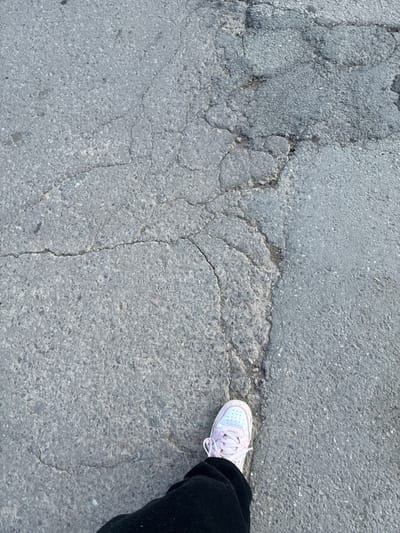 The scene depicts a damaged asphalt surface in Madrets, Bulgaria, viewed from an overhead perspective. A human left foot, covered by a light pink and white sneaker with white laces, is positioned on a substantial longitudinal crack in the asphalt. The visible portion of the leg is clad in dark-colored trousers. The asphalt exhibits extensive deterioration, including numerous intersecting cracks, fissures, and areas of aggregate exposure across the entire visible ground plane.