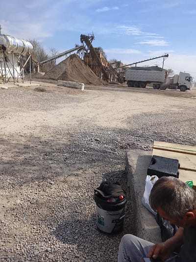 The image depicts an industrial sand and gravel processing plant located in the city of Yangi Chinoz, Uzbekistan, under a partially cloudy blue sky. In the lower right foreground, a male individual is seated on a concrete curb, facing away from the camera, revealing his greyish-brown hair, dark shirt, and blue jeans. His hands are visible, appearing to manipulate a small electronic device. Adjacent to him on the gravel ground is a black and grey bucket labeled "LONGMAN," containing dark fabric material. On the concrete curb, next to the seated person, are a white plastic bag, a black rectangular container, and several wooden planks.
The ground surface across the scene is primarily light-colored gravel and dirt, with visible shadows. In the midground, a prominent conical pile of dark brown aggregate material is present. Behind this pile, the industrial plant consists of various interconnected conveyor belts, elevated structures, and processing machinery. To the far left, several large, silver-insulated cylindrical tanks are mounted on white support structures. To the right, a white heavy-duty semi-trailer dump truck is positioned, its trailer covered with a light green/grey tarp, suggesting it is either being loaded or waiting. A long, rectangular white concrete block lies on the ground between the aggregate pile and the truck. Sparse, bare trees are visible in the distant background.