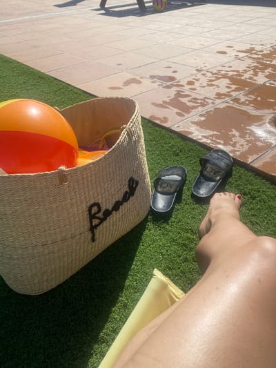 A woman's tanned legs are visible in the foreground, with one foot, painted with red nail polish, resting near a pair of black slides on artificial turf. Next to the slides, a woven straw beach bag is positioned, spilling out an orange and yellow inflatable ball. The bag has the word "Beach" spelled out in black rope attached to its side. In the background, a tiled patio area is wet and sun-dappled, suggesting recent activity near a pool. The overall scene evokes a relaxed, sunny day outdoors, likely in Spain, given the location context. The bright sunlight and the presence of beach items indicate it's daytime and the weather is warm.