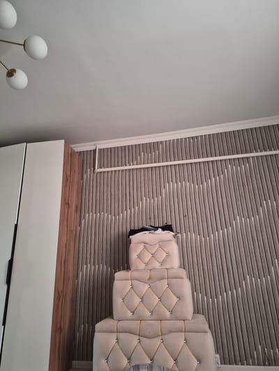 The image depicts an interior scene with a focus on a decorative wall and furniture. On the left, a section of a tall white wardrobe with a wooden accent is visible. To the right of the wardrobe, a three-tiered piece of upholstered furniture, likely a headboard or ottoman, is positioned against a wall covered in a textured wallpaper. The wallpaper features a pattern of vertical, light-colored slats with darker recessed areas, creating a rhythmic, wave-like effect. The upholstered furniture is a light beige color and features diamond-shaped tufting with gold cord trim and silver studs at the intersections. A few dark items of clothing are draped over the top tier of the furniture. In the upper left corner, a modern light fixture with multiple white globe shades is partially visible against a white ceiling. The image appears to be taken from a low angle, looking upwards. The scene is clean and organized, suggesting a residential or hotel setting. There is no indication of the city of Bereke, Kazakhstan, within the image itself.