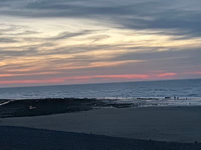 This image depicts a serene beach scene at dusk in Veules-les-Roses, France. The sky is painted with soft hues of pink, orange, and grey, with wispy clouds stretching horizontally across the horizon. The sea is calm, with gentle waves lapping at the shore. A dark, rocky section of the beach is visible to the left, partially submerged by the receding tide, revealing pools of water. Further to the right, a sandy stretch of the beach extends towards the water, where a few small figures are seen standing near the water's edge. The foreground is dominated by a dark, pebbly beach. The overall atmosphere is peaceful and tranquil, characteristic of a quiet evening by the sea.