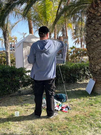 A person in a blue shirt and black pants stands painting on an easel among palm trees in Germasogeia, Cyprus. The artist wears a black hat. The scene is sunlit with shadows on the grass. A white reflective board is propped up next to the artist, and painting supplies are scattered on the ground nearby. A partially visible beach is in the background.