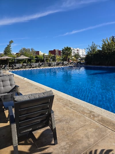The image depicts an outdoor swimming pool area in Taza, Morocco, under a clear blue sky with sparse wispy clouds. The foreground features light brown tiled paving with shadows from unseen foliage, on which a dark gray metal chair with gray cushions is positioned with its back to the viewer, next to a small glass-topped table and another similar chair. The rectangular swimming pool, filled with bright blue water, occupies the midground. Along the far side of the pool, numerous dark gray cushioned lounge chairs are arranged, interspersed with several light beige parasols and green trees and shrubs. In the background, multi-story buildings of various colors, including red and white, are visible, confirming an urban setting. No individuals are present in the scene.