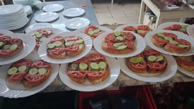 In the city of Marchevo, Bulgaria, a kitchen scene unfolds with numerous plates of open-faced sandwiches prepared and ready for serving. The sandwiches are topped with slices of tomato and cucumber, arranged in a way that suggests a smiley face on some. These culinary creations are presented on white, decorative plates, stacked and arranged across the countertops.

Adjacent to the prepared food, stacks of clean white plates are visible, hinting at a larger event or a busy food service operation. The countertops themselves are a mix of metallic and tiled surfaces, reflecting the utilitarian nature of a kitchen environment. Further in the background, a rustic wooden table, likely used for food preparation or service, adds to the overall rustic aesthetic of the space. The overall impression is one of a well-organized kitchen preparing for a meal service, likely for a communal gathering or event within Marchevo.