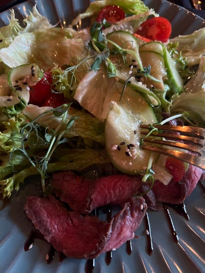 The image displays a close-up, overhead view of a salad served on a dark, ridged plate. The salad contains sliced cucumbers, cherry tomatoes, lettuce, microgreens, and thinly sliced rare beef. A fork is embedded in a slice of cucumber, with sesame seeds scattered throughout the salad. A dark, glossy sauce is drizzled on the plate. The lighting suggests an indoor setting, possibly a restaurant, with reflections visible on the salad ingredients. The overall impression is of a fresh and well-prepared dish. No people, text, or specific time of day or weather cues are visible. The context provided indicates the location is Kopanskoi, Russia.