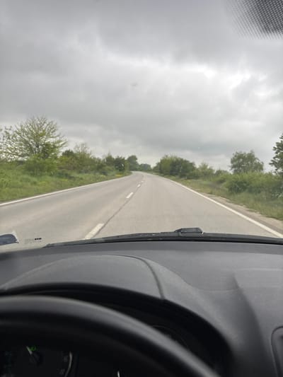 The image presents a view from the interior of a vehicle, looking out onto a long, asphalt road that stretches into the distance and appears to curve slightly to the left. A solid white line marks the right edge of the road, while a dashed white line is visible on the left side, likely indicating the center or passing lane.
The road is flanked on both sides by green vegetation, consisting of trees, bushes, and some grassy areas, suggesting a rural or semi-rural setting within Trilistnik, Bulgaria. Above, the sky is heavily overcast with dense grey clouds, indicating a cloudy or potentially gloomy day with diffused light.
In the foreground, on the right side, elements of the vehicle's interior are visible. These include a grey dashboard, the lower portion of the windshield, and a windshield wiper blade positioned upright. Part of a steering wheel or a circular gauge can be seen in the bottom right, with partial numerical markings such as "50" and "55". A sticker on the windshield displays fragmented text, including "ABC D-EV". The overall impression is one of a journey or drive through the countryside on an overcast day.